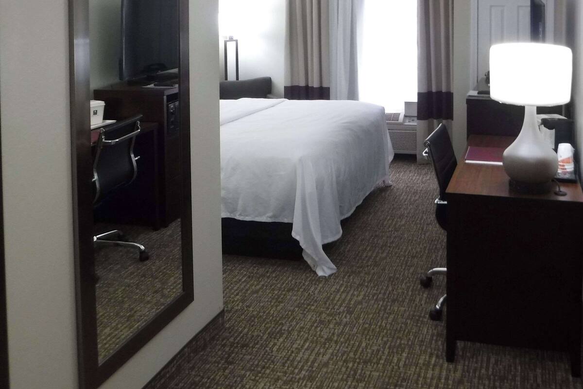 A hotel room with a bed, a desk, a chair, a lamp, and a large mirror.