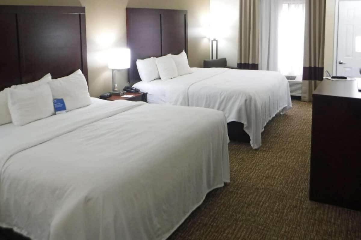 A hotel room with two beds, a desk, a lamp, and a view of the outdoors.