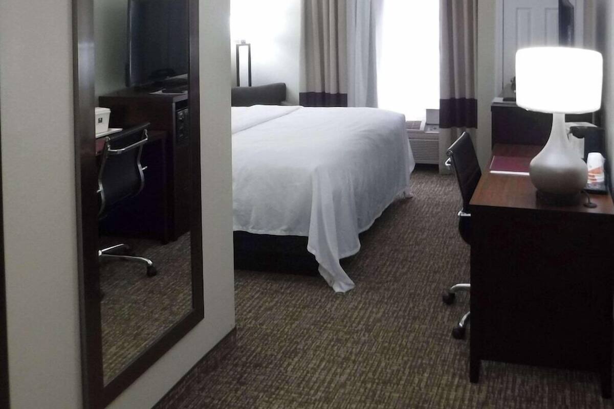 A hotel room with a bed, a desk, a chair, a lamp, a mirror, and a door.