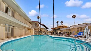 A swimming pool with a clear blue water, surrounded by lounge chairs and umbrellas, and a building with balconies and palm trees in the background. - Super 8 by Wyndham Los Angeles-Culver City Area (Los Angeles)