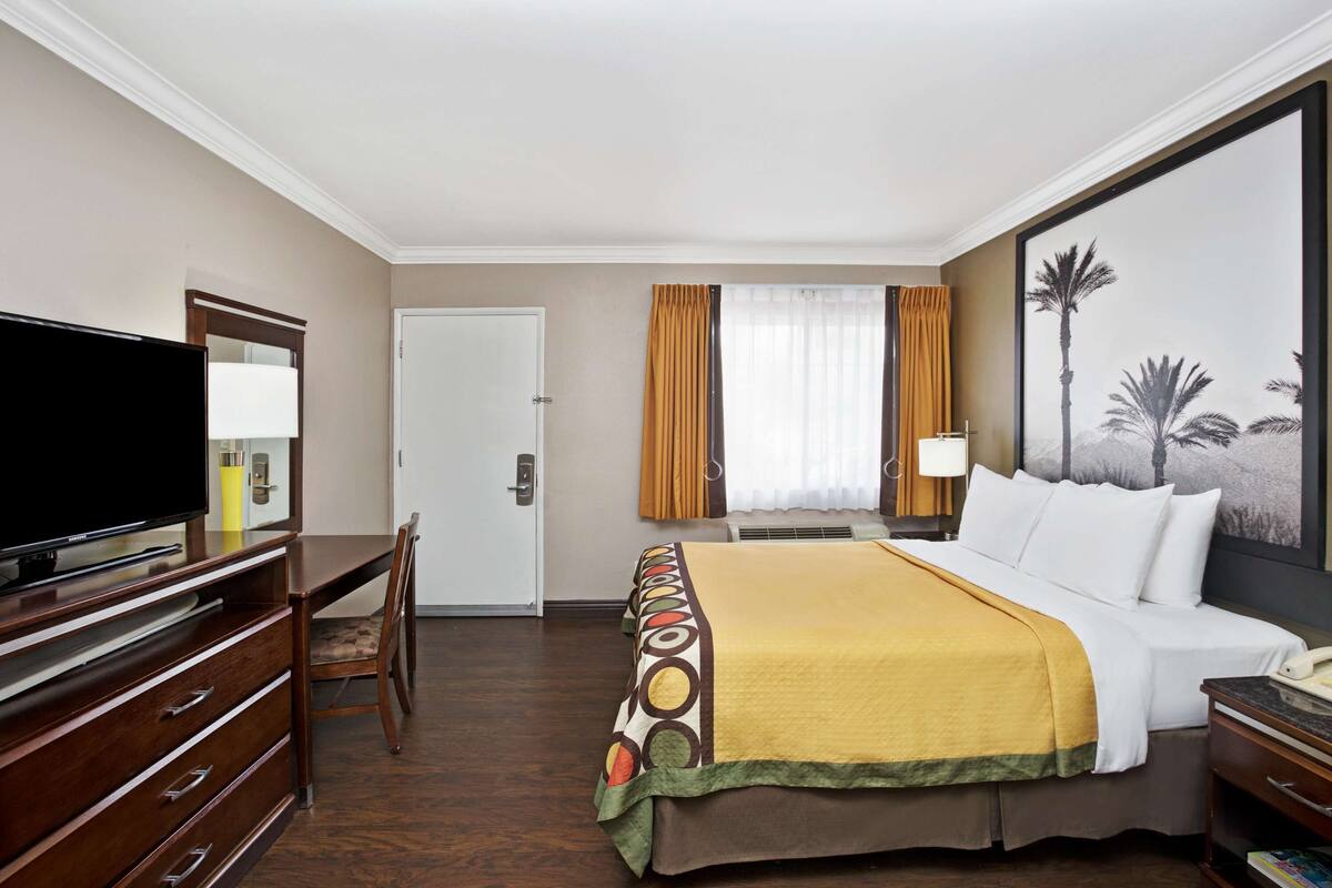 Une chambre d’hôtel comprenant un lit, un bureau avec une télévision, une chaise, une fenêtre avec des rideaux et un tableau au mur.