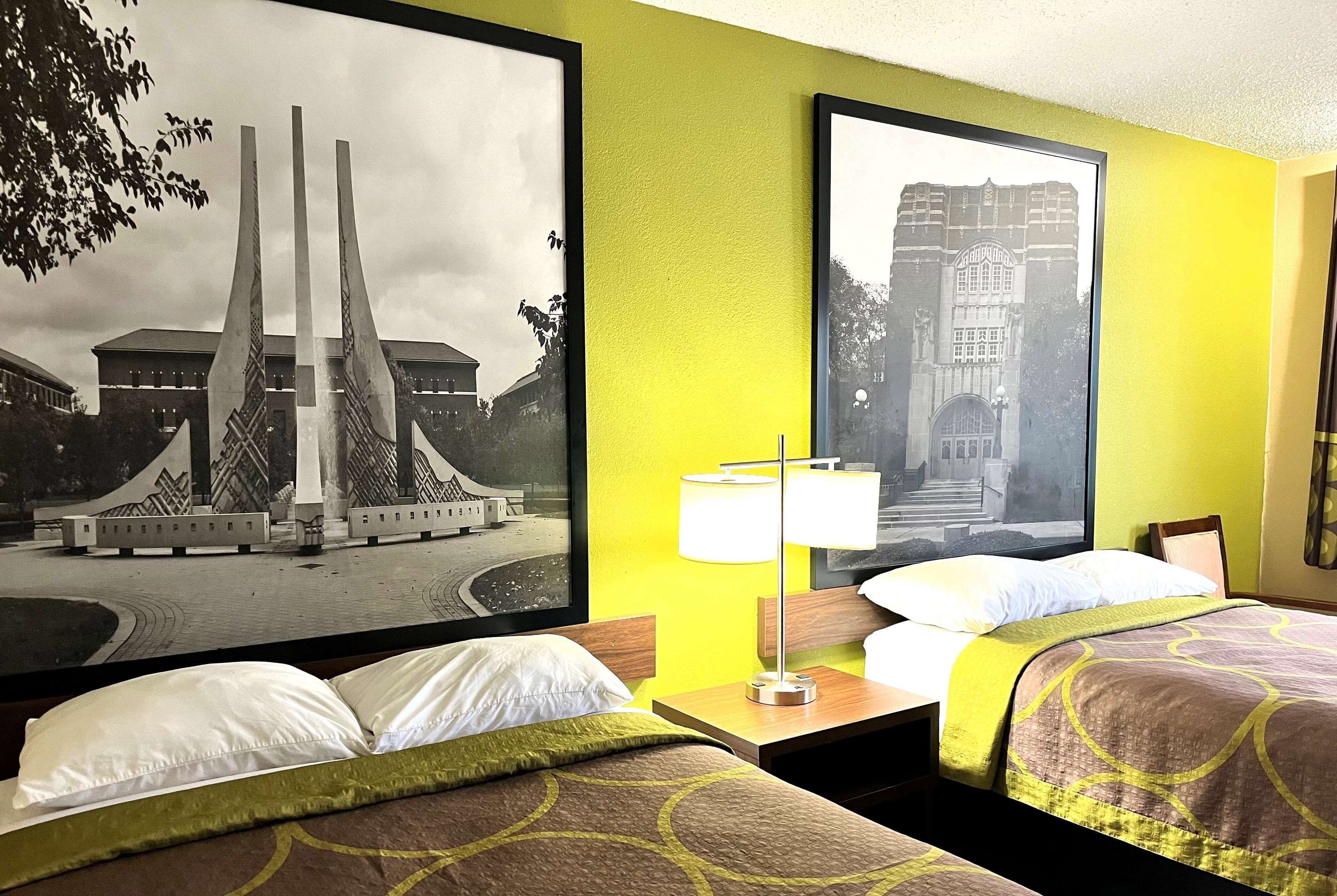 Quarto de hotel com duas camas, uma mesa de cabeceira com abajur e duas grandes molduras na parede.