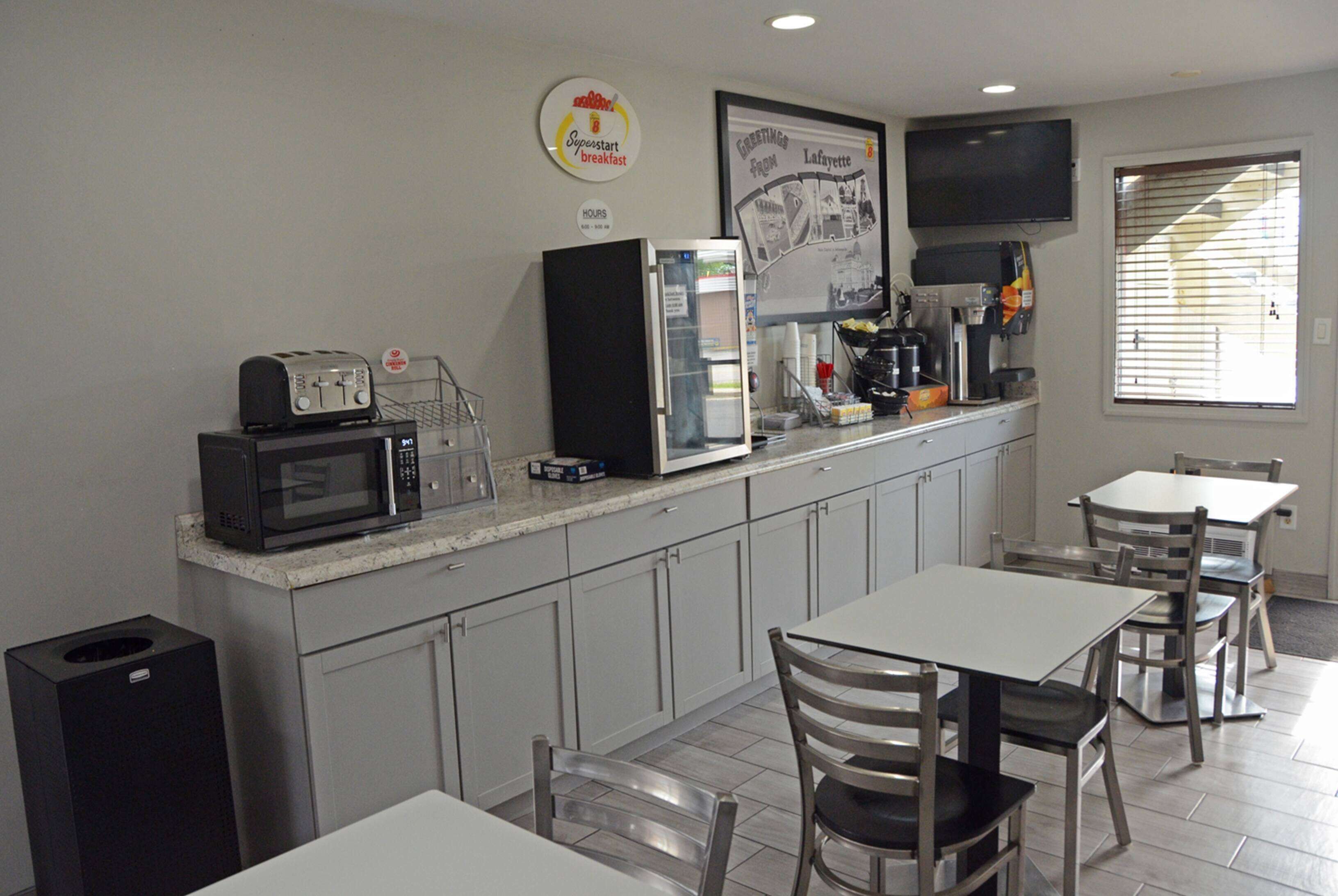 A kitchenette with a microwave, toaster, refrigerator, coffee maker, and a coffee station. There are two tables and chairs, and a framed picture on the wall.