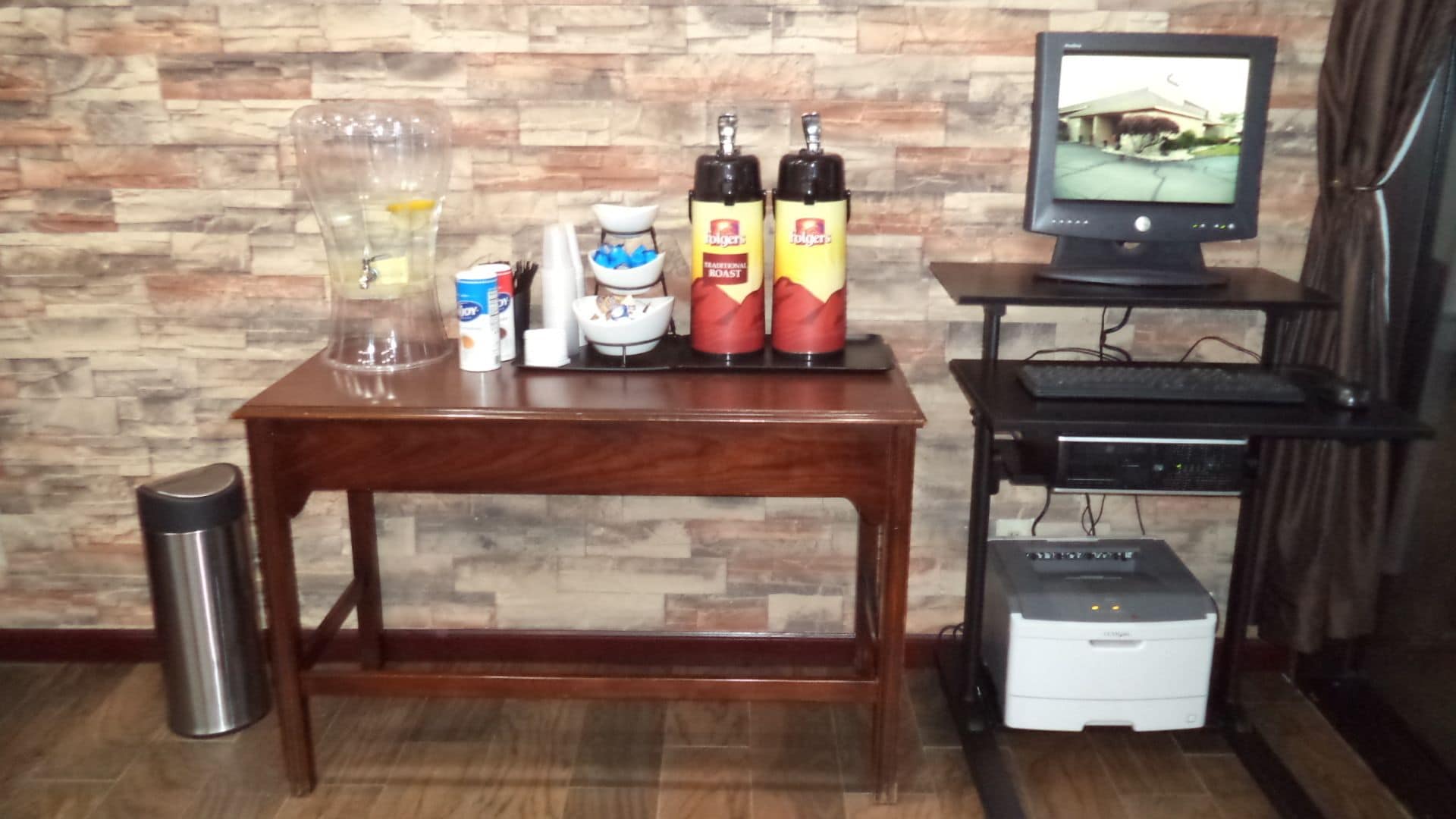 Una mesa de madera con un recipiente de vidrio, dos botellas, un cuenco pequeño y una lata. Una impresora está sobre un soporte junto a la mesa.