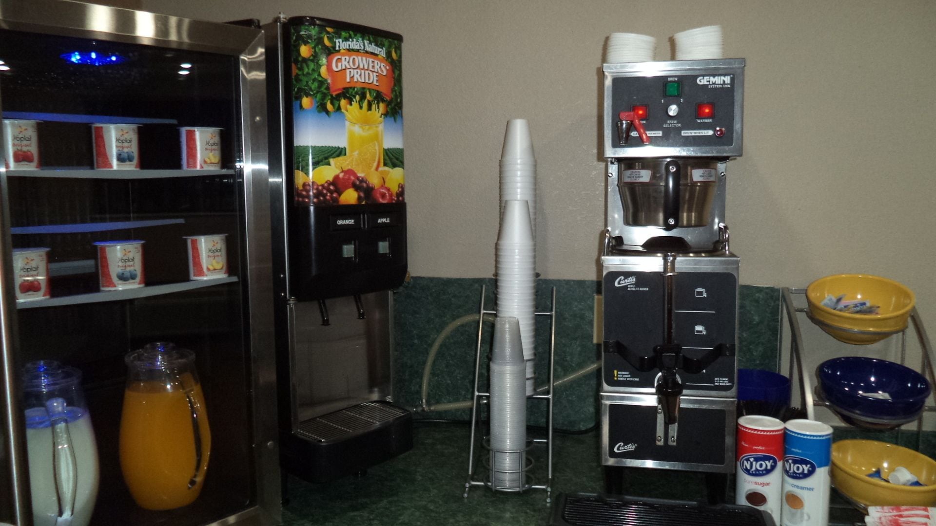 A self-serve coffee station with a beverage dispenser, a juice dispenser, and a selection of condiments and utensils.