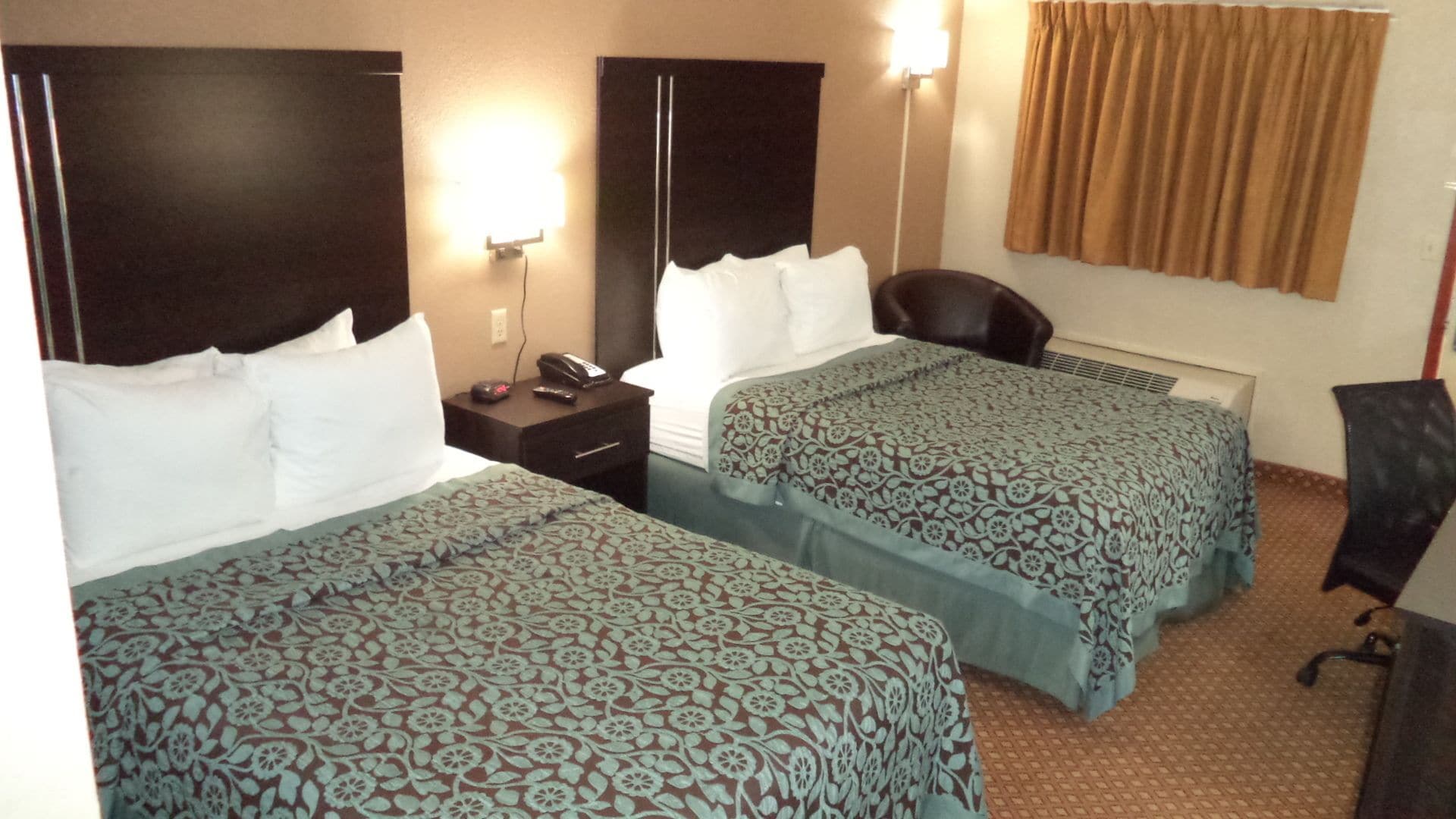 A hotel room with two beds, a desk, a chair, and a lamp.