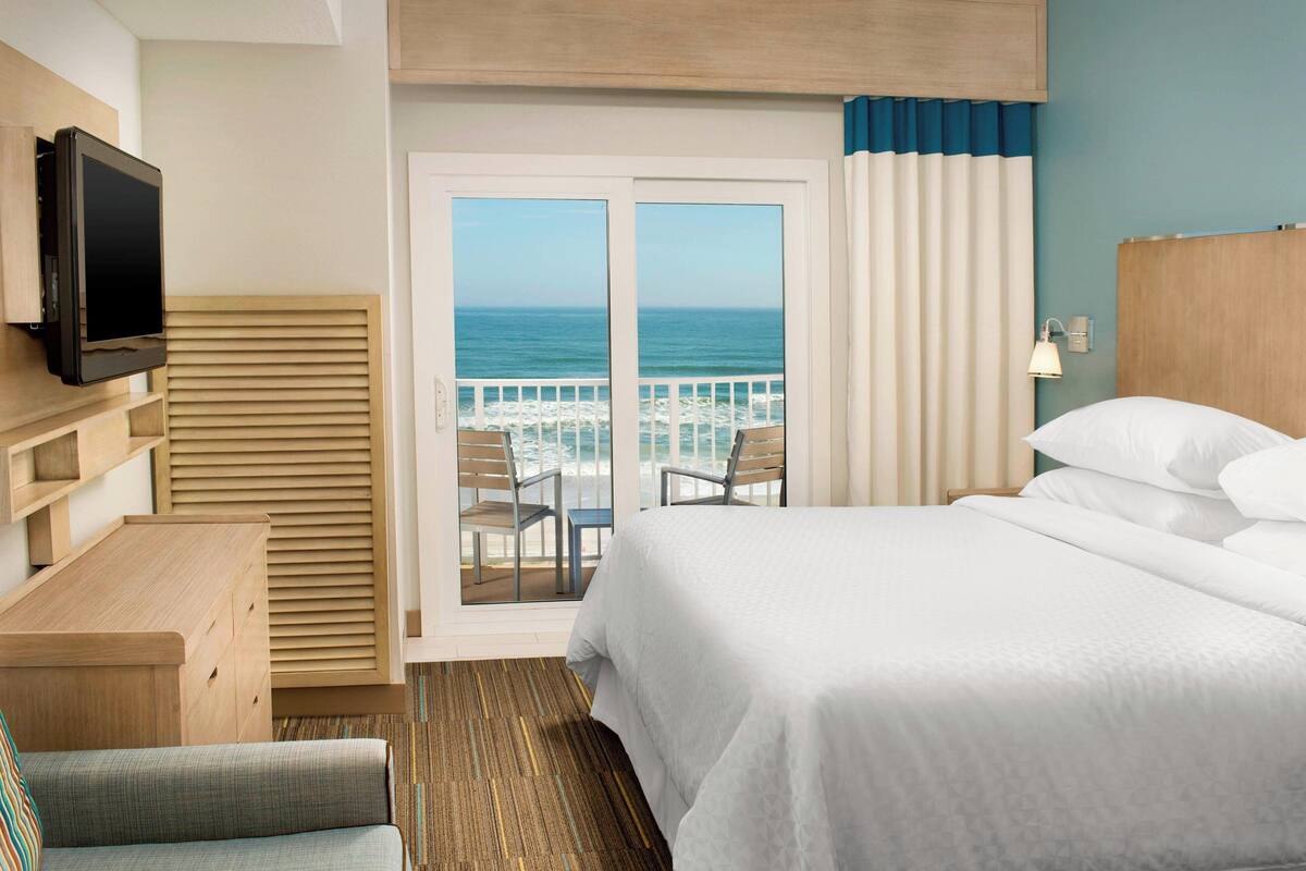 Habitación de hotel con cama, televisión, escritorio y balcón con vista al océano.