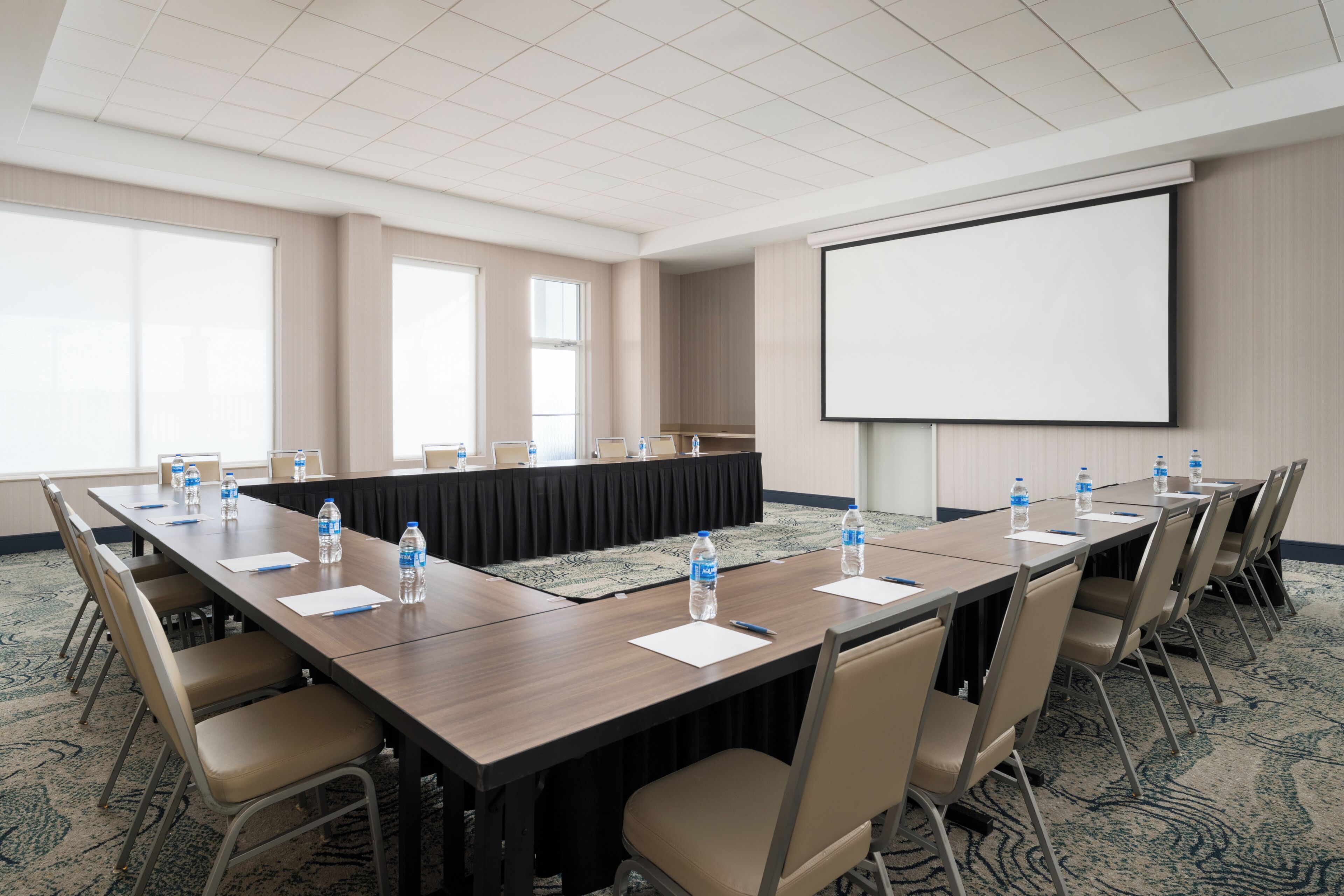 Un salón de conferencias con una larga mesa, sillas, botellas de agua y una pantalla de proyector.