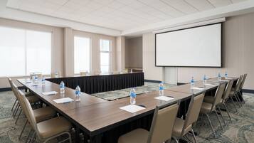 Un salón de conferencias con una larga mesa, sillas, botellas de agua y una pantalla de proyector.