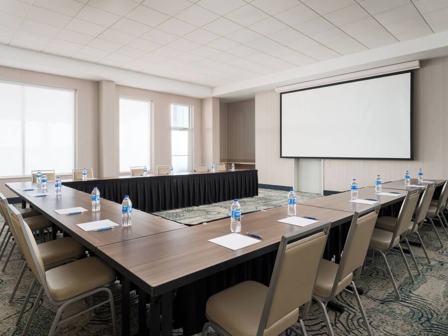 Un salón de conferencias con una larga mesa, sillas, botellas de agua y una pantalla de proyector.
