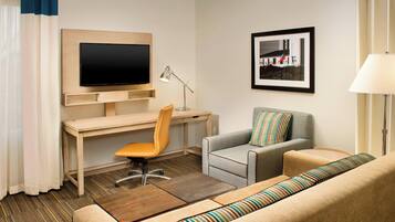 A hotel room with a wooden desk, a television, a grey sofa, a striped sofa, a wooden coffee table, and a lamp.