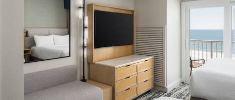 Habitación de hotel con cama, televisor, cómoda de madera, espejo y vista al mar.