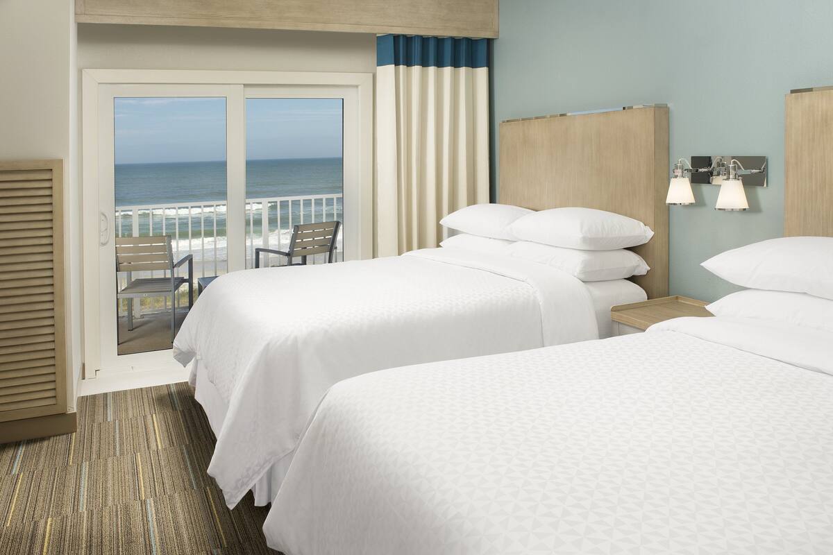 Habitación de hotel con dos camas, vista al océano, balcón con una silla y cabecera de madera.