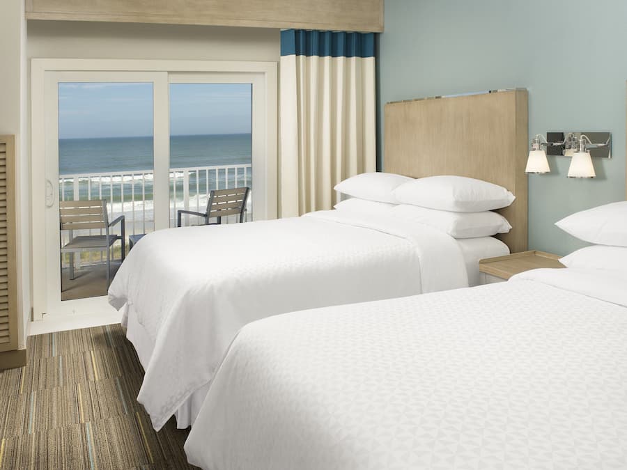 A hotel room with two beds, a view of the ocean, a balcony with a chair, and a wooden headboard.