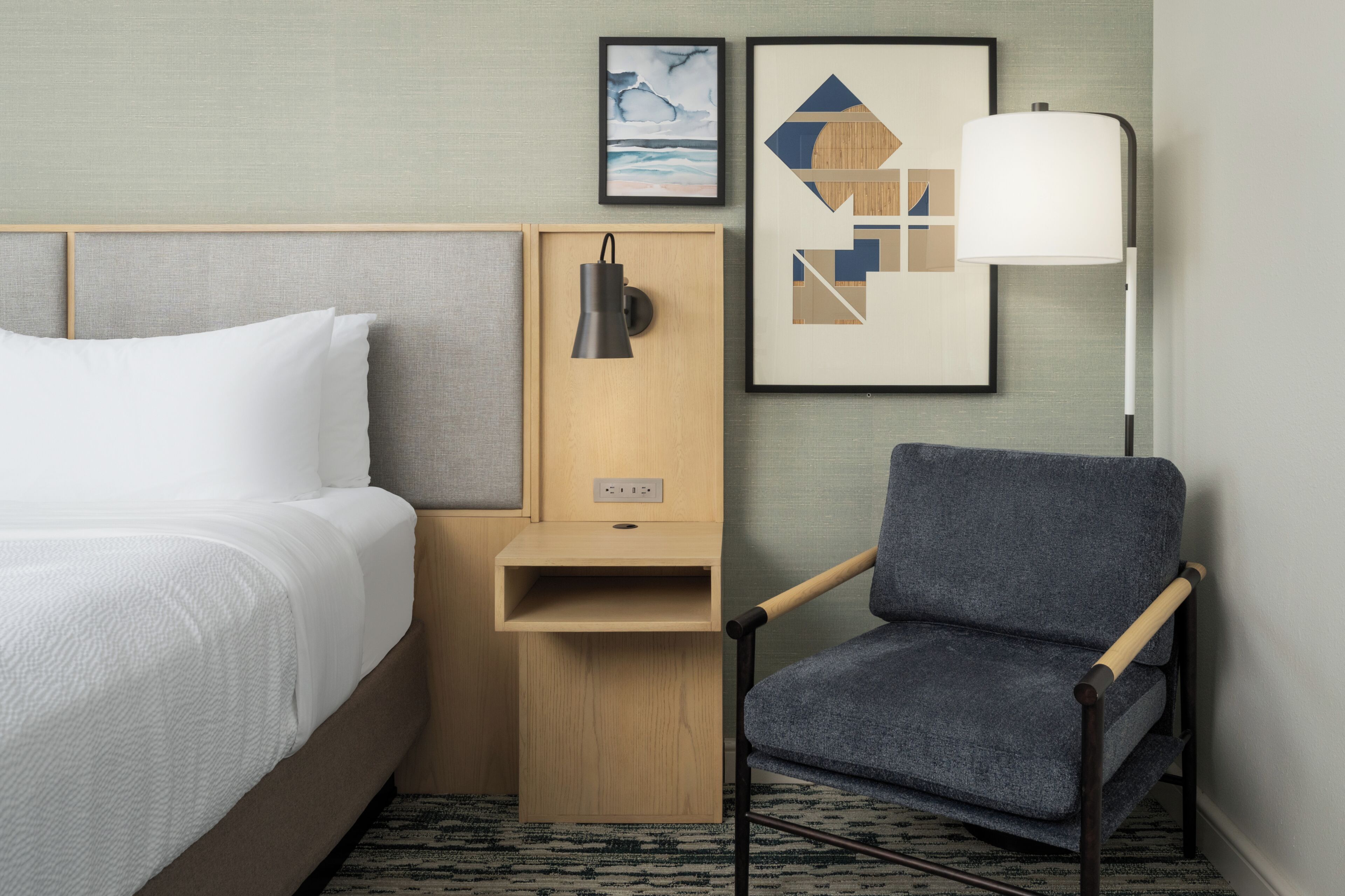 Ein Hotelzimmer mit einem Bett, einem Sessel, einem Nachttisch, einer Lampe und zwei gerahmten Bildern an der Wand.