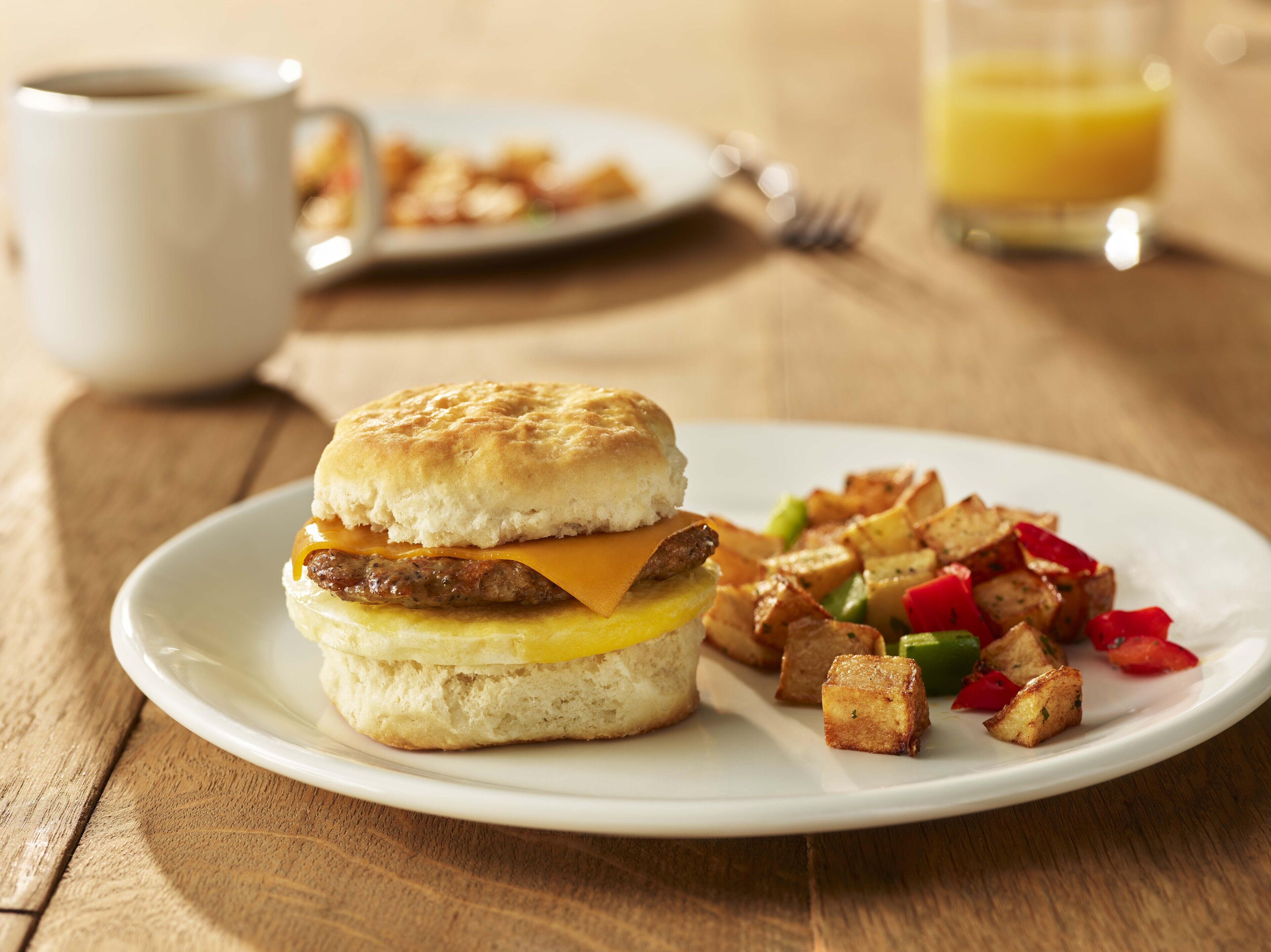 A breakfast plate with a sausage sandwich, hash browns, and a glass of orange juice.