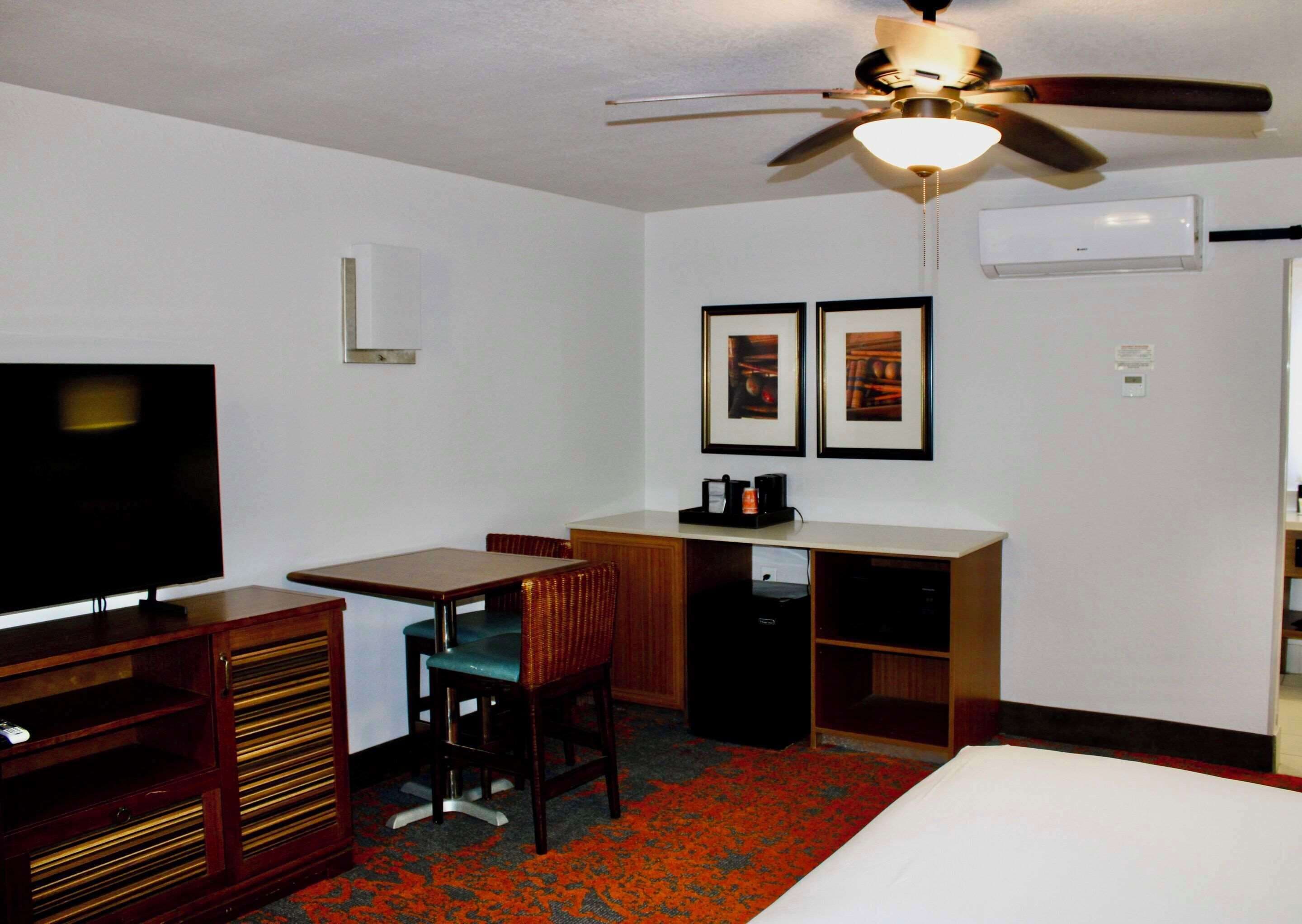A hotel room with a bed, a desk with a chair, a television, a ceiling fan, and framed pictures on the wall.