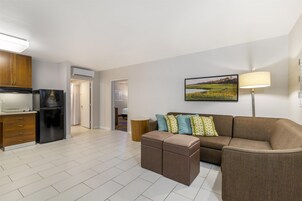 Una sala de estar moderna con sofá, reposapiés y una lámpara. Cuenta con una zona de cocina con refrigerador y alacenas. En la pared hay un cuadro, y un equipo de aire acondicionado está instalado.