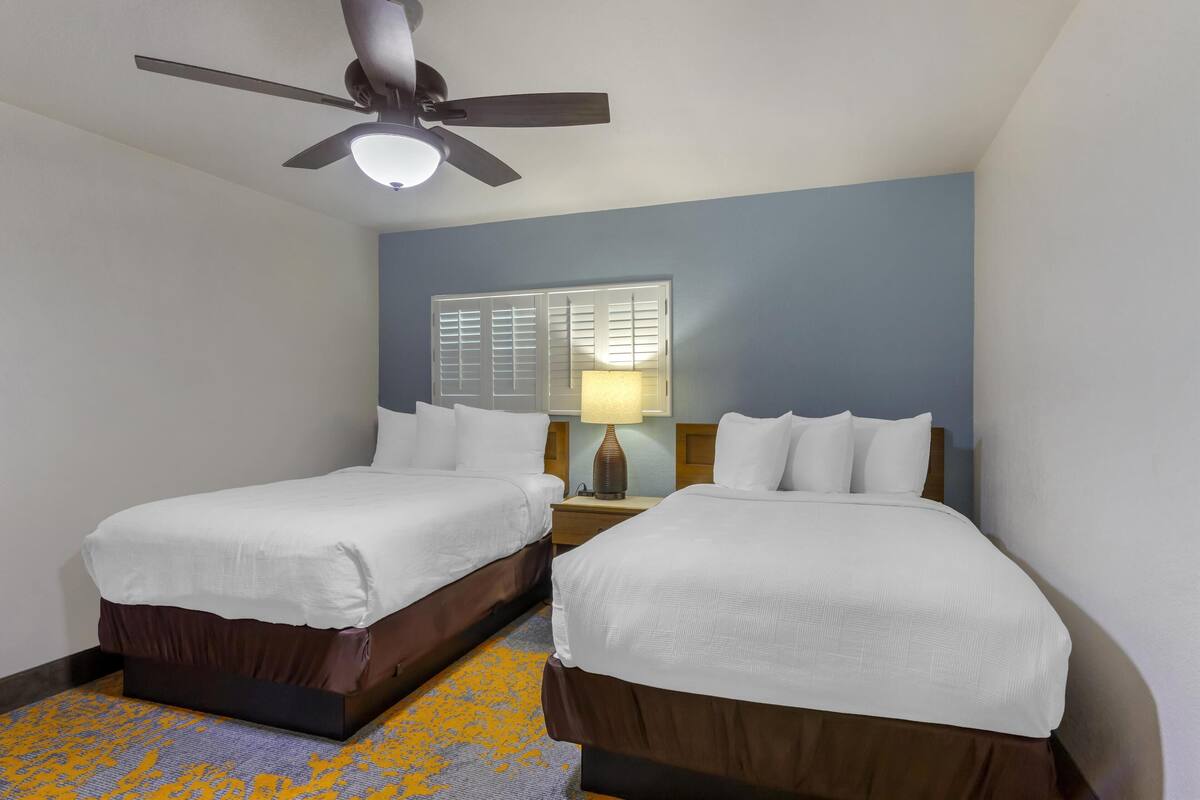 A hotel room with two single beds, a ceiling fan, a window with blinds, and a lamp on the nightstand.