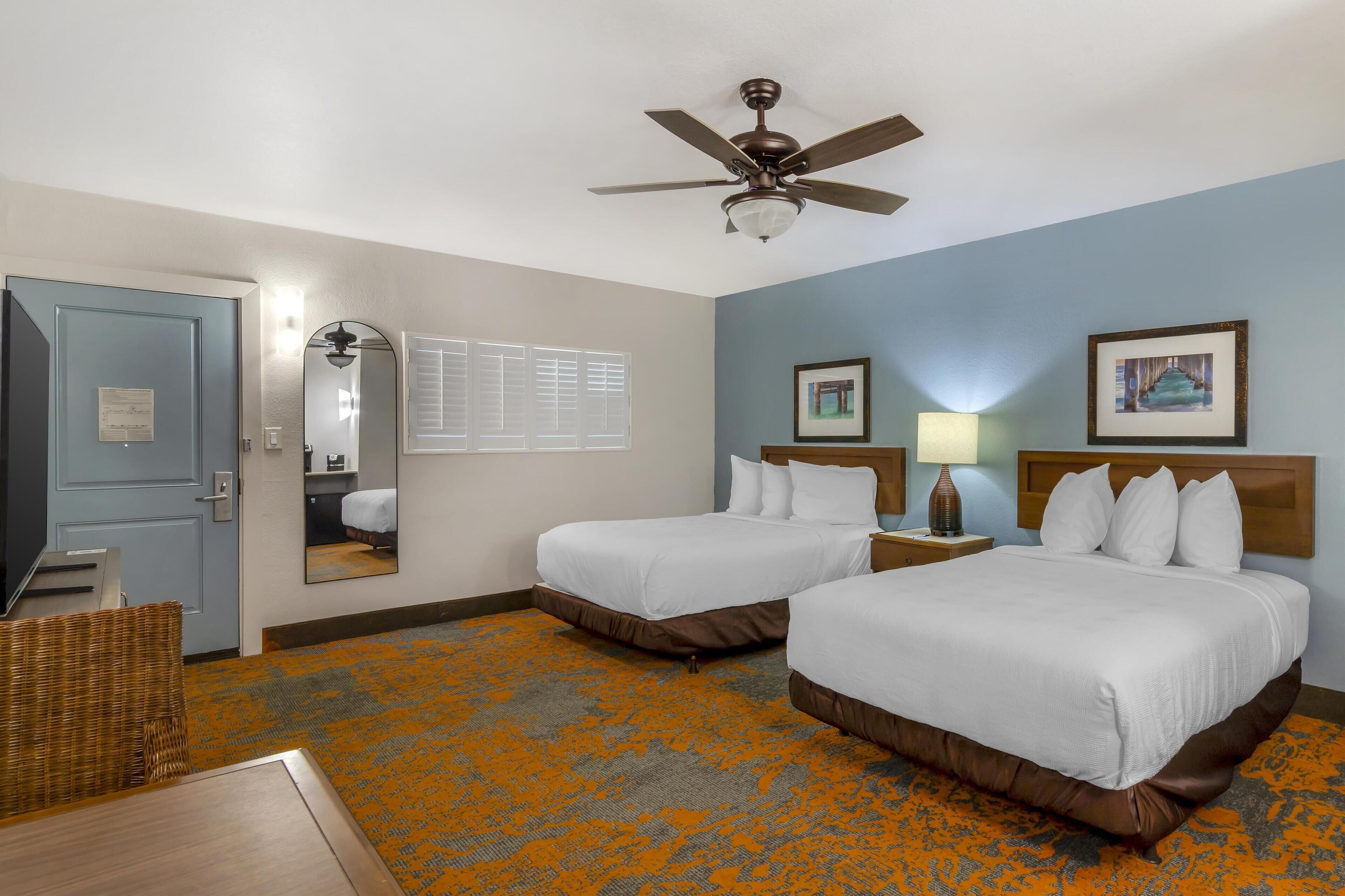 A hotel room with two beds, a ceiling fan, a television, a mirror, and a window with blinds.
