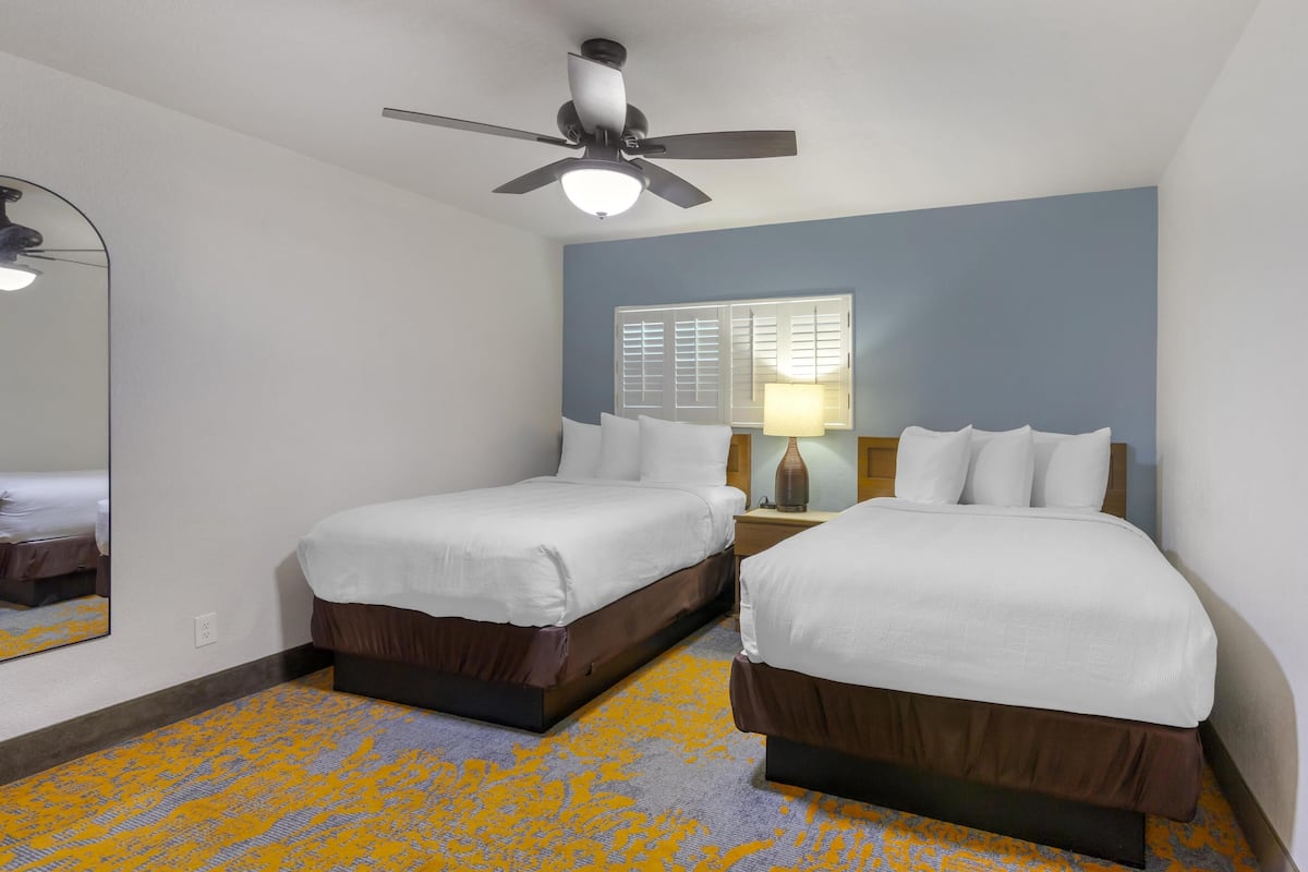 A hotel room with two beds, a ceiling fan, a window with blinds, a mirror, and a patterned carpet.