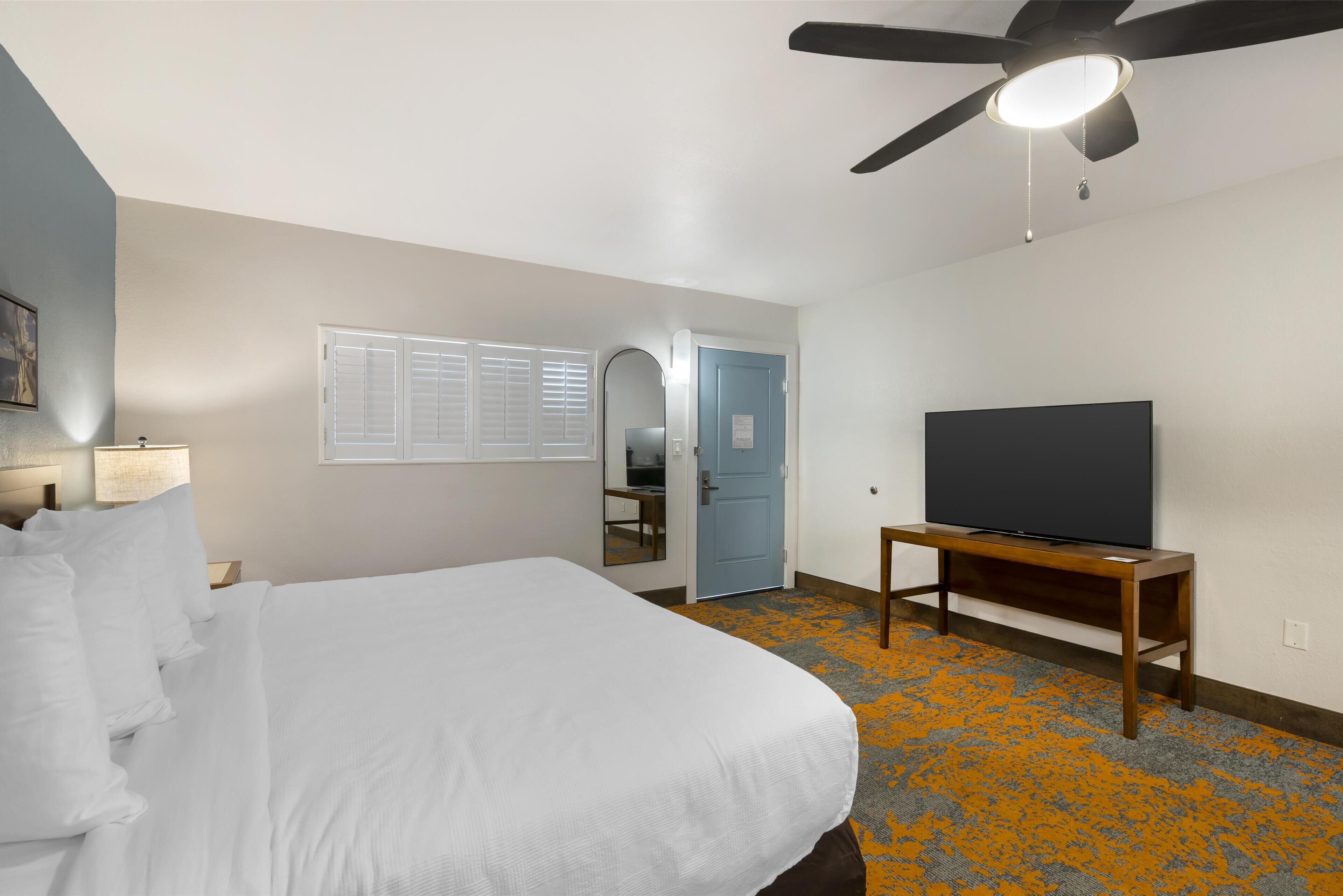 A hotel room with a bed, a television on a stand, a ceiling fan, and a door leading to a bathroom.