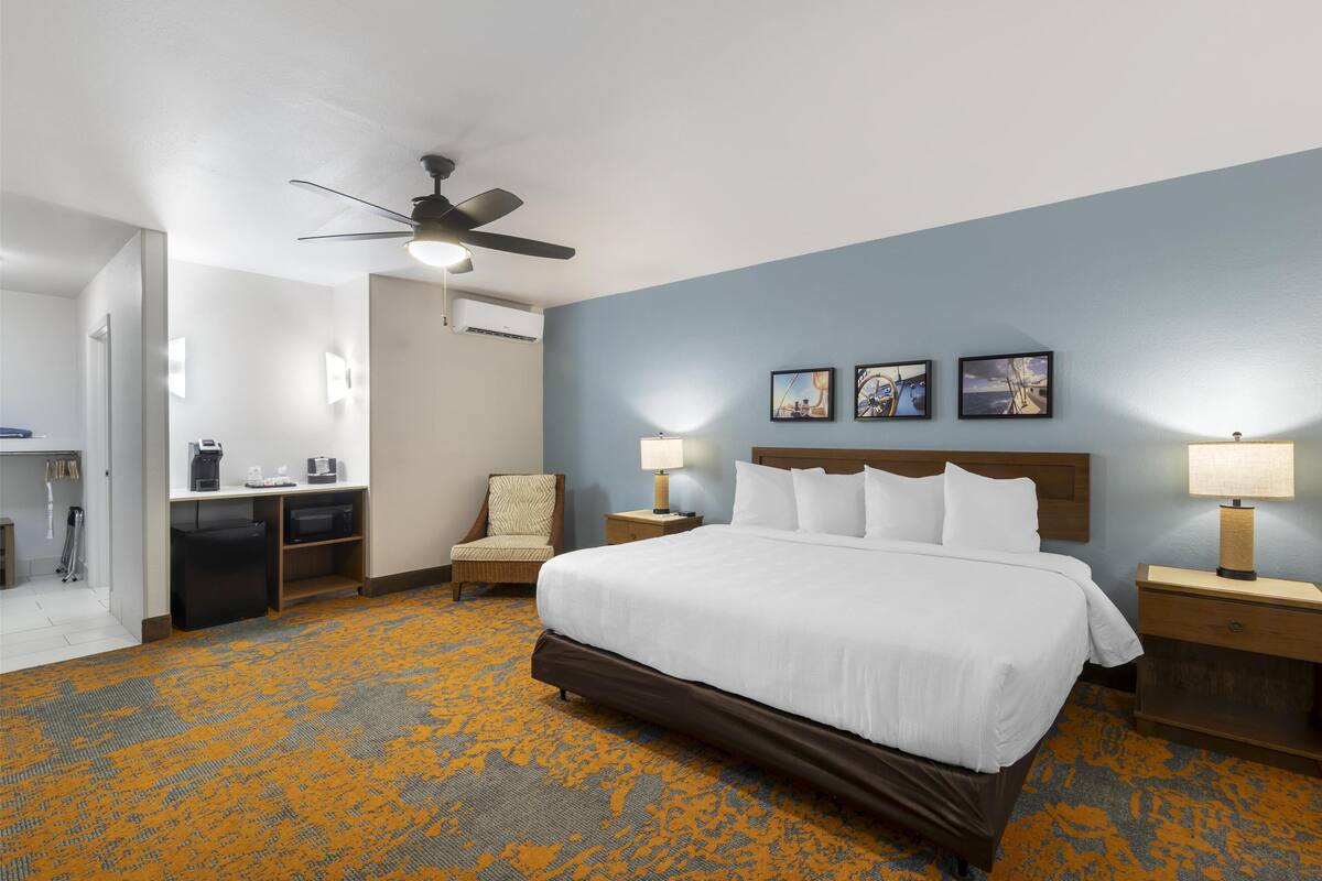 A hotel room with a bed, a desk, a chair, a ceiling fan, and a wall with three framed pictures.