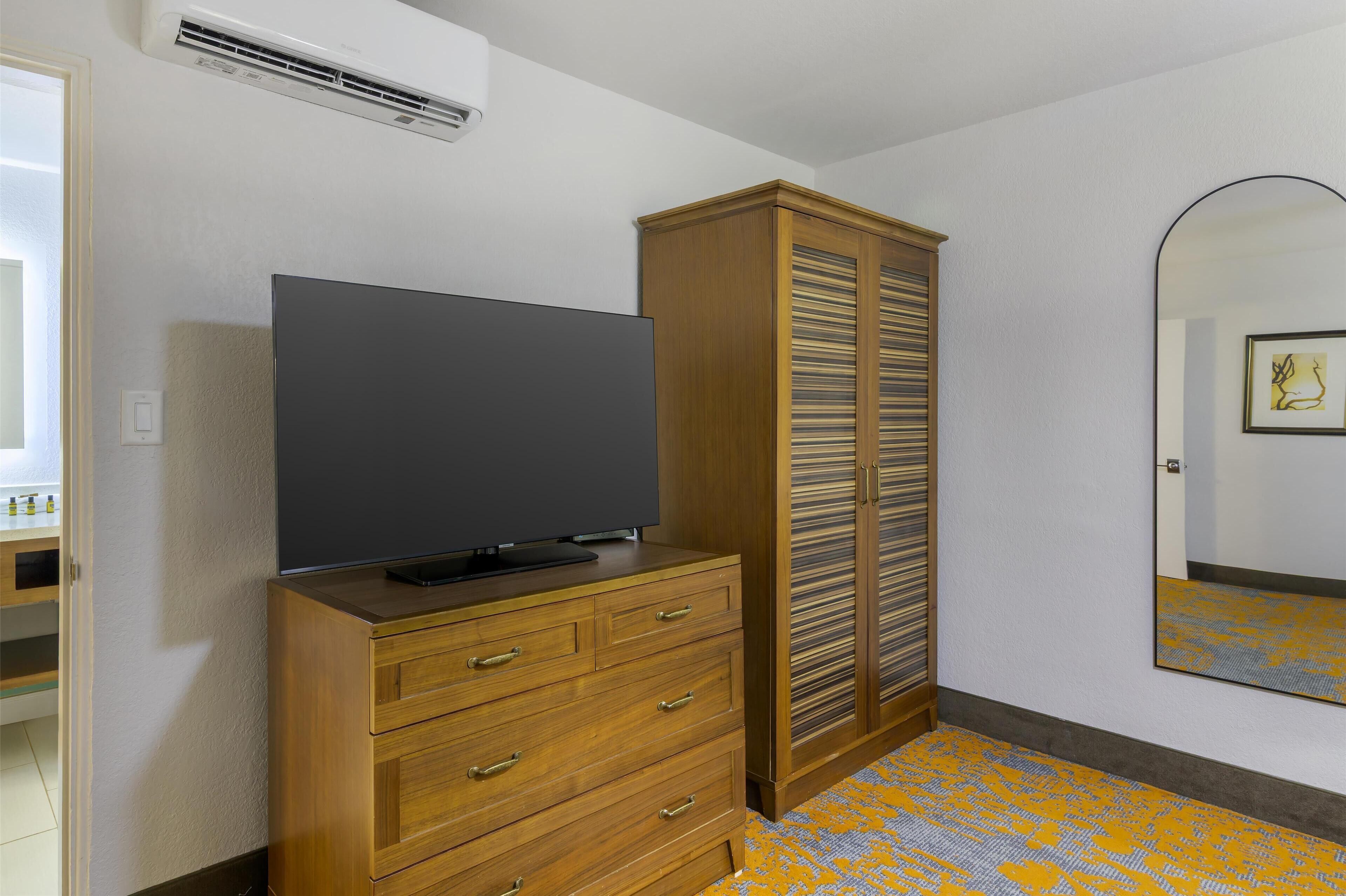 A room with a wooden dresser, a flat-screen TV, a wardrobe, and a mirror on the wall.