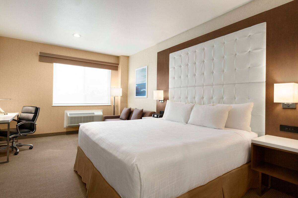 A hotel room with a large bed, a desk with a chair, a window with blinds, and a wall-mounted lamp.