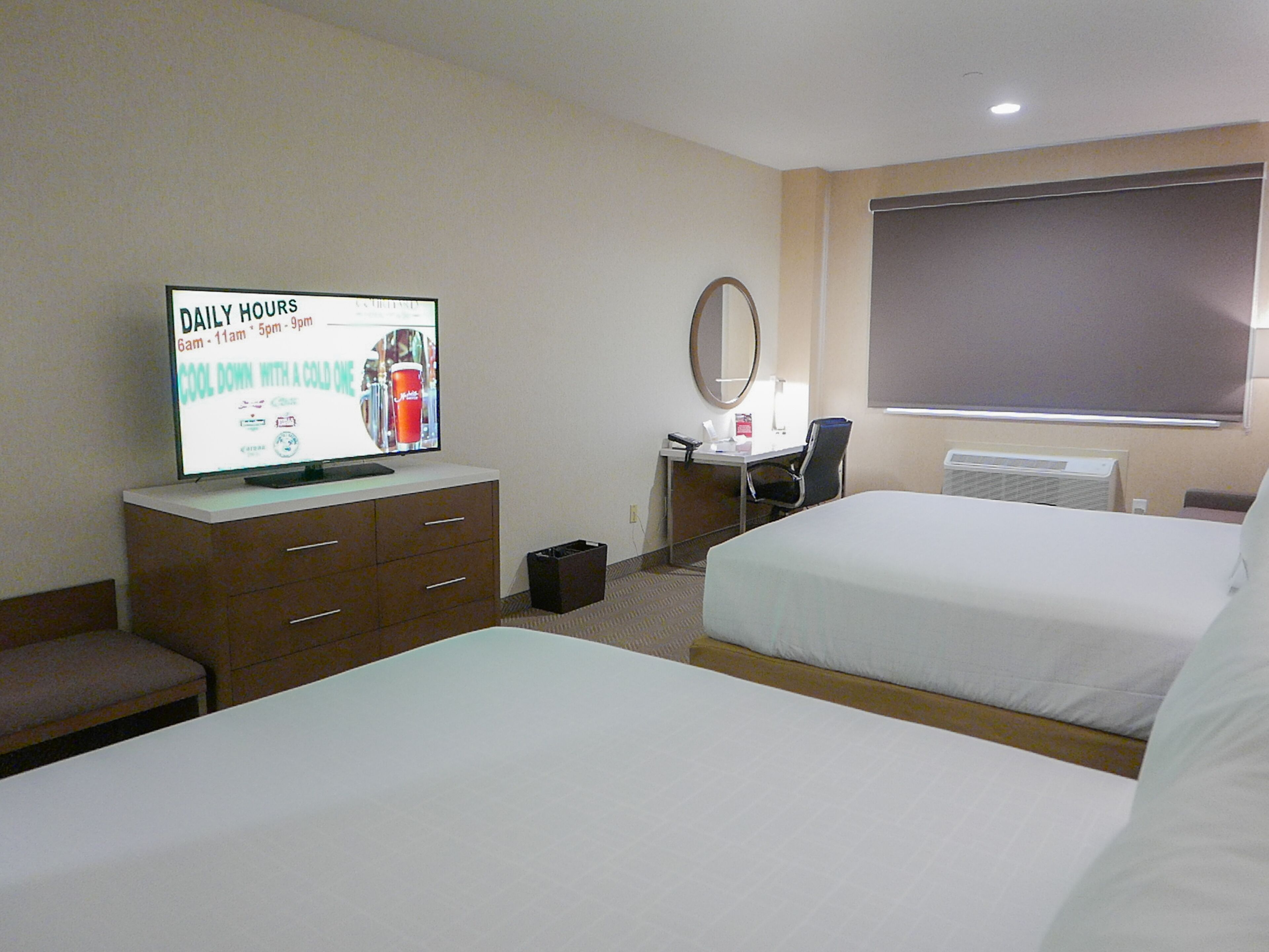 A hotel room with two beds, a desk with a computer, a television displaying daily hours, and a large window.