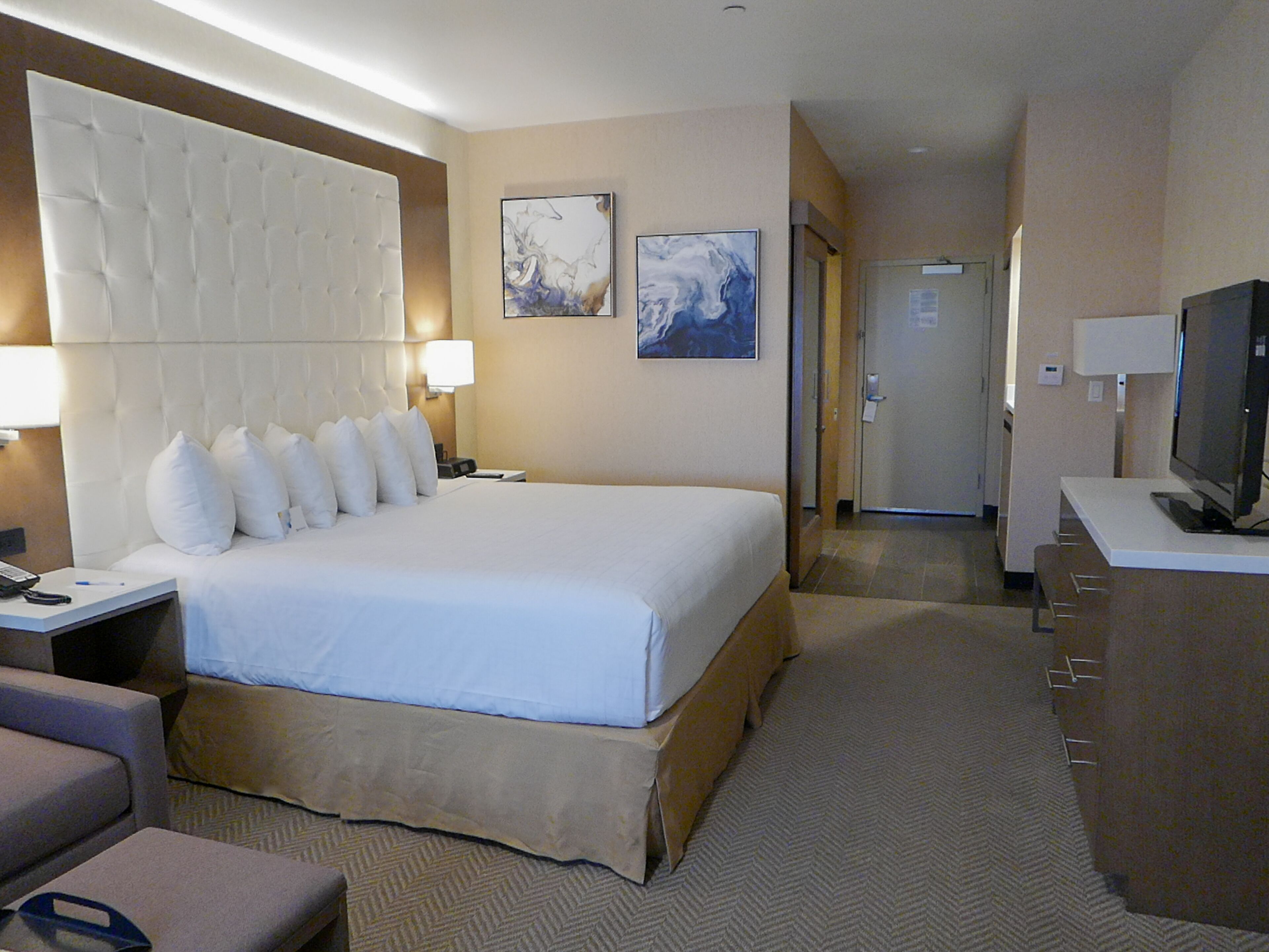 A hotel room with a large bed, a desk with a computer, a TV, and two artwork pieces on the wall.