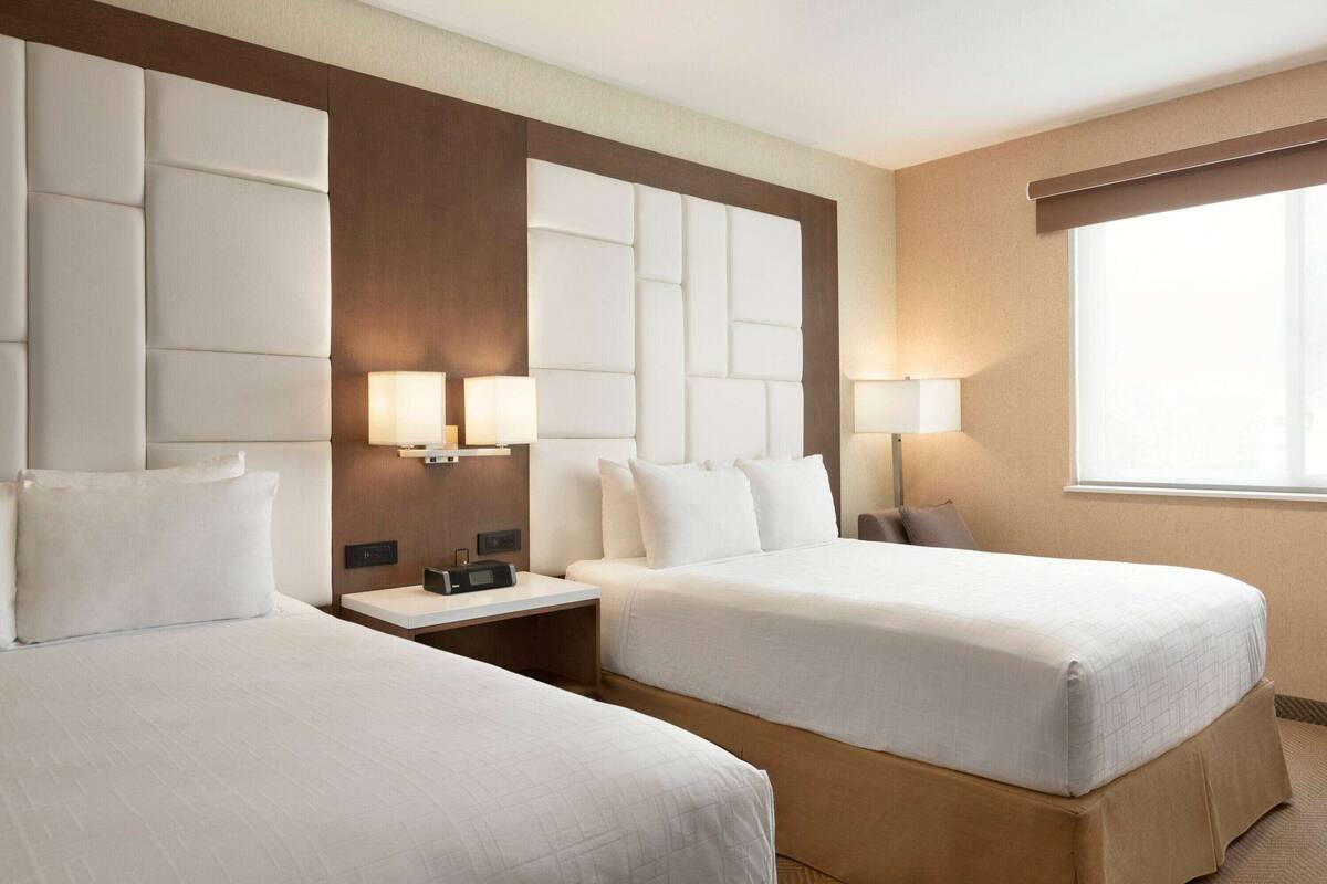 A hotel room with two beds, a wooden headboard, bedside lamps, and a large window.
