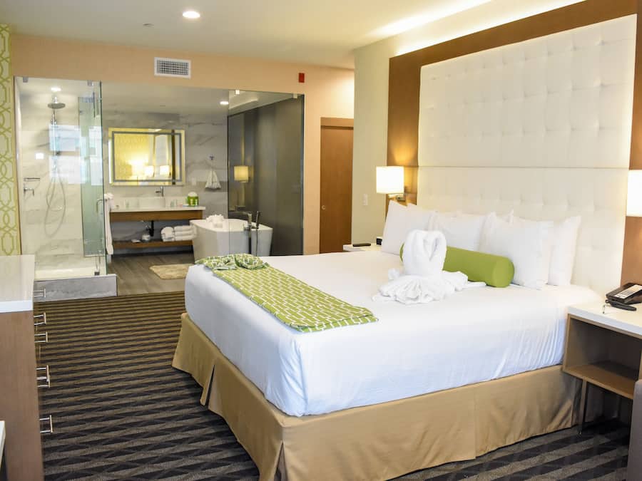 A hotel room with a large bed, a separate bathroom with a glass shower, a bedside table with a lamp, and a small desk with a telephone.