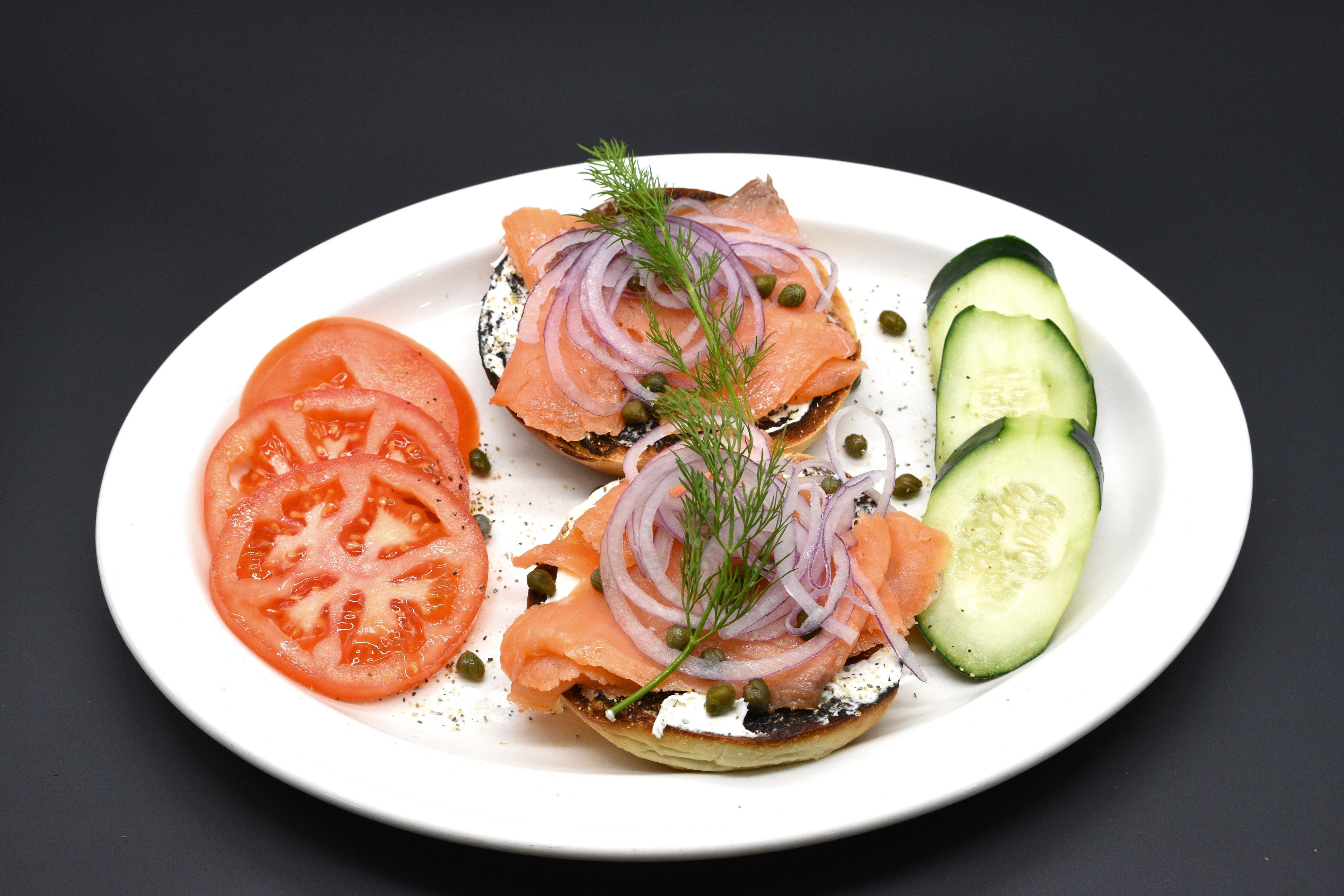 A plate with bagels topped with smoked salmon, red onion, capers, and dill, accompanied by slices of tomato and cucumber.