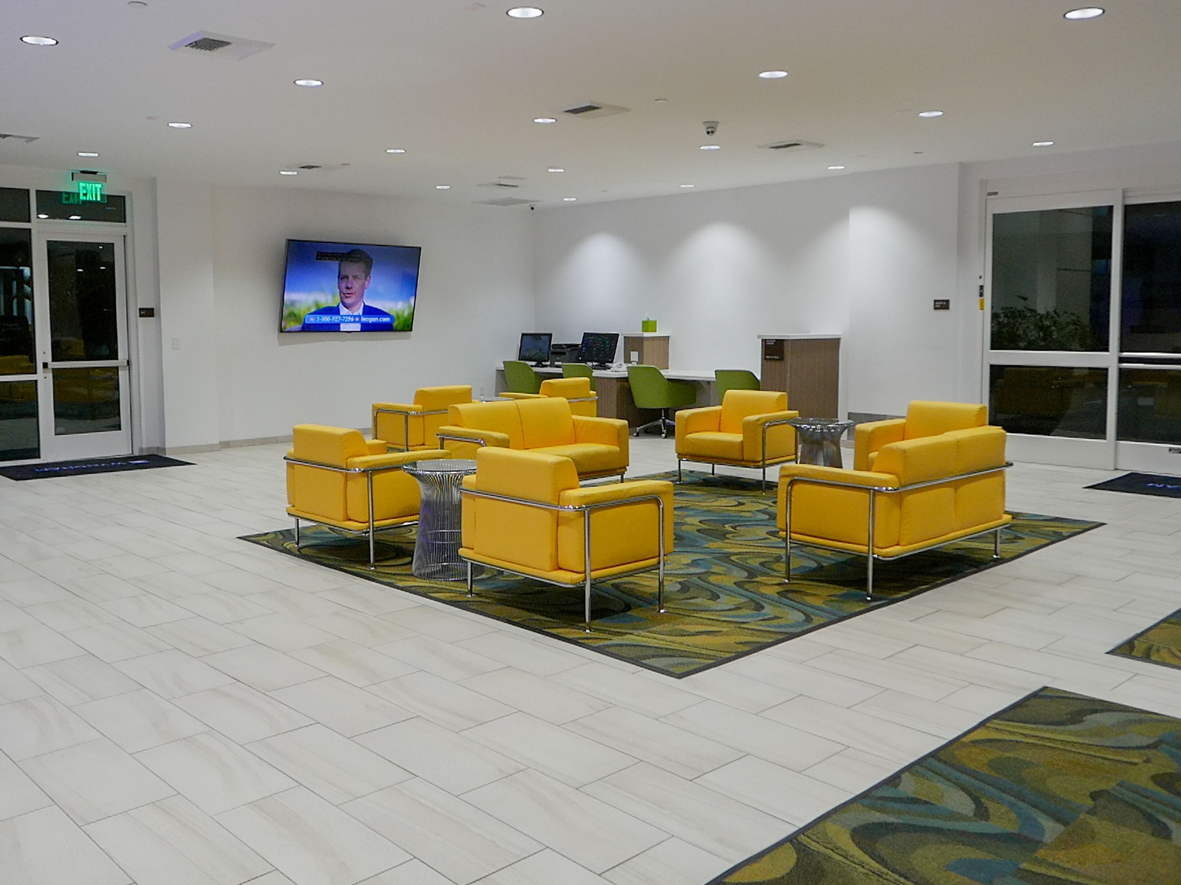A lobby with yellow chairs arranged in a semi-circle, a flat-screen TV on the wall, and a reception desk with computers.