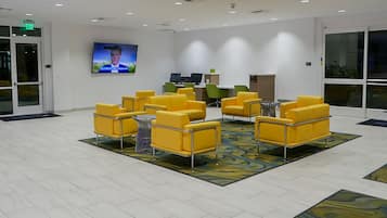 A lobby with yellow chairs arranged in a semi-circle, a flat-screen TV on the wall, and a reception desk with computers.