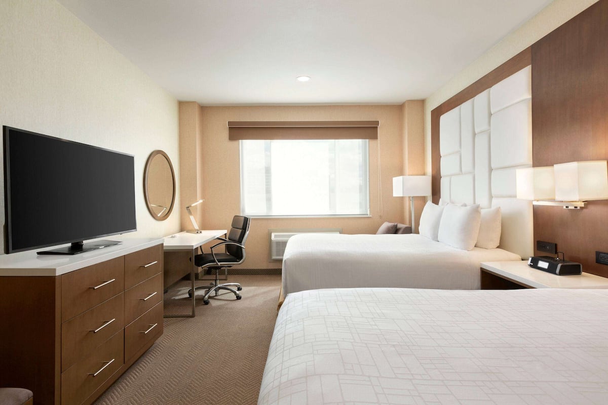 A hotel room with two beds, a desk with a chair, a flat-screen TV, a mirror, and a large window with blinds.