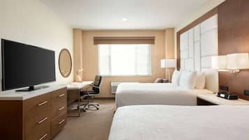 A hotel room with two beds, a desk with a chair, a flat-screen TV, a mirror, and a large window with blinds.