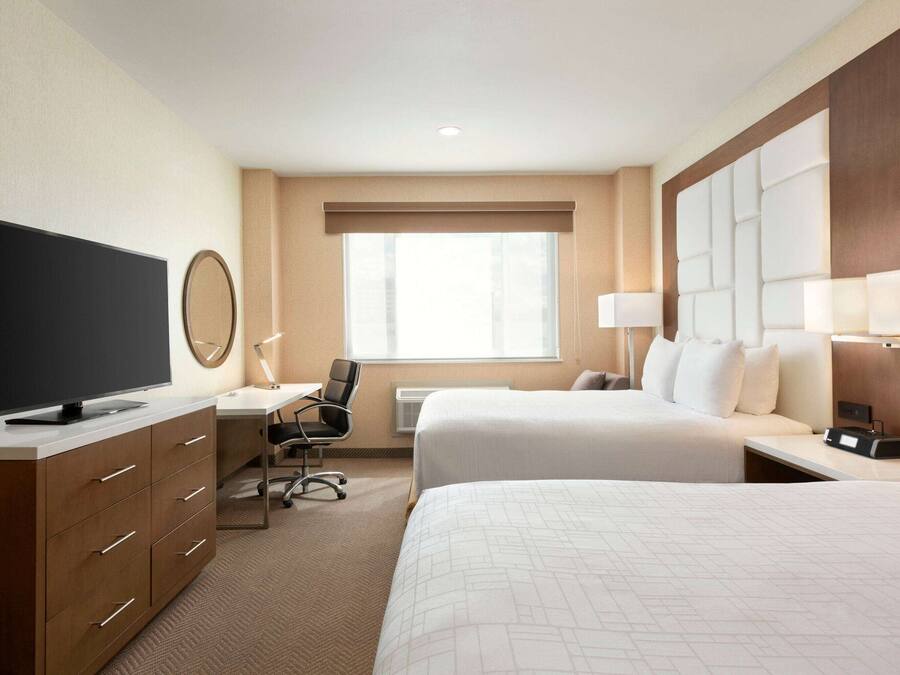 A hotel room with two beds, a desk with a chair, a flat-screen TV, a mirror, and a large window with blinds.