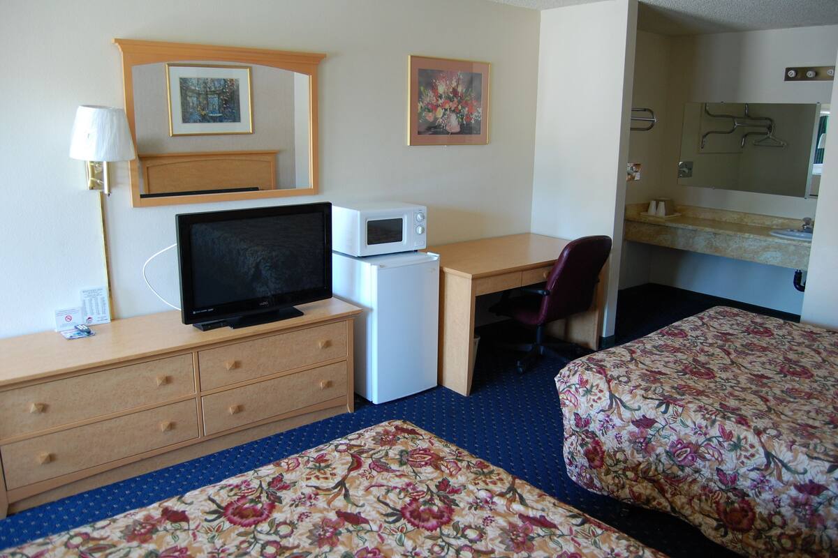 Ein Hotelzimmer mit einem Bett, einem Schreibtisch mit Computer, einer Mikrowelle, einem Kühlschrank, einem Fernseher, einer Lampe und einem durch eine offene Tür einsehbaren Badezimmer.