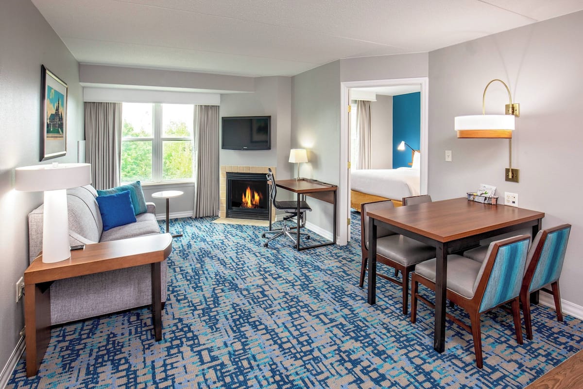 A hotel room with a fireplace, a sofa, a dining table with chairs, a TV mounted on the wall, and a view of the outdoors.