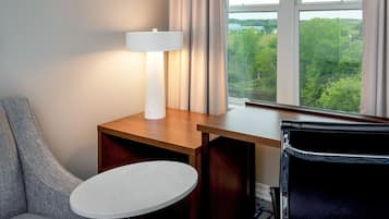 A hotel room with a wooden desk, a round table, a grey armchair, and a view of greenery through the window.