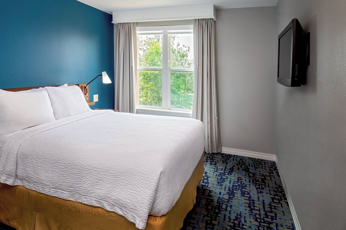 A hotel room with a bed, a window with curtains, a television on the wall, and a patterned carpet.