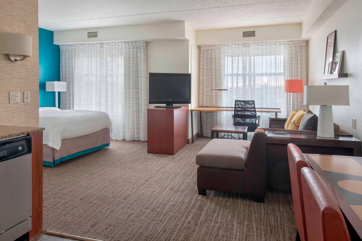 A hotel room with a bed, a desk with a chair, a television, and a dining table with chairs.