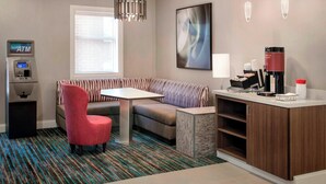 Habitación de hotel con sofá, una mesita, una silla roja, un cajero automático, una cafetera y un ventanal grande.