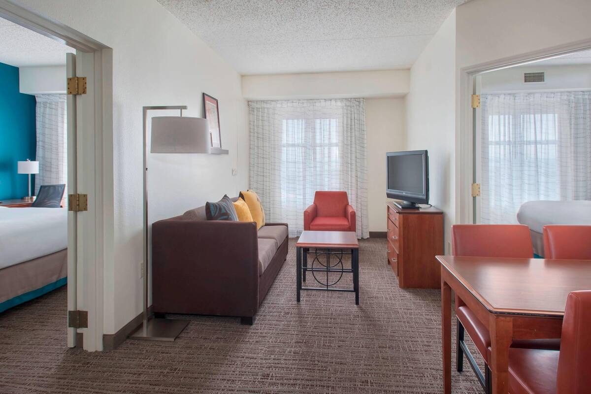 A hotel room with a bed, a sofa, a red armchair, a TV, and a dining table.