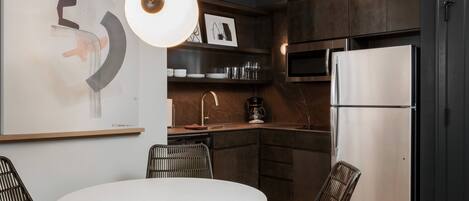 A modern kitchen with dark cabinetry, a stainless steel refrigerator, a white dining table, and wicker chairs.