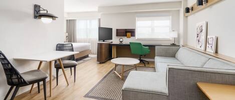 Una habitación de hotel moderna con sofá, una mesa de comedor con sillas, una mesita redonda, televisor y un escritorio con computadora.