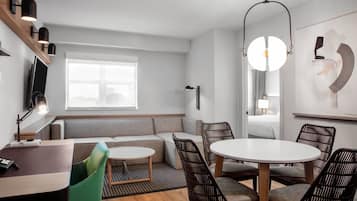A modern hotel room with a sofa, dining table, and chairs, a television, and a large window providing natural light.