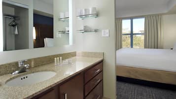 A hotel bathroom with a granite countertop sink, a mirror, and glass shelves above the sink holding towels.